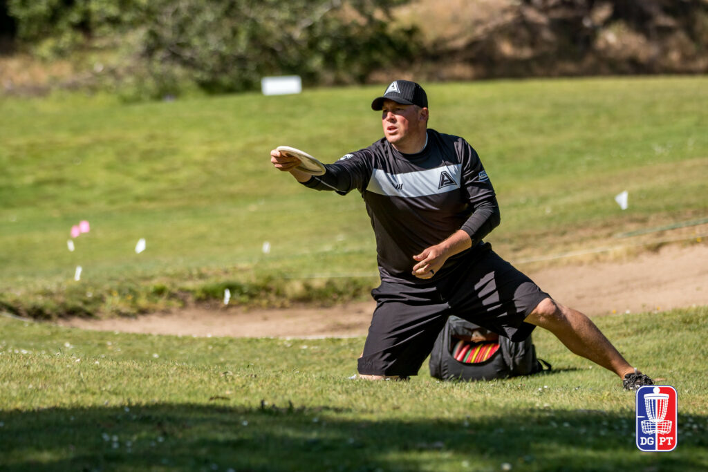 Avery Jenkins kneels in the grass while lining up a putt. His GRIPeq disc golf bag sits on the ground behind him.