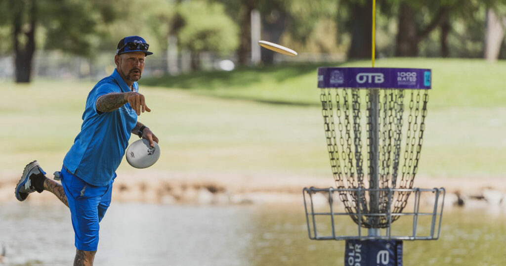 Patrick Brown putting into a disc golf basket. The disc is mid-flight and his leg is outstretched from the putting motion.