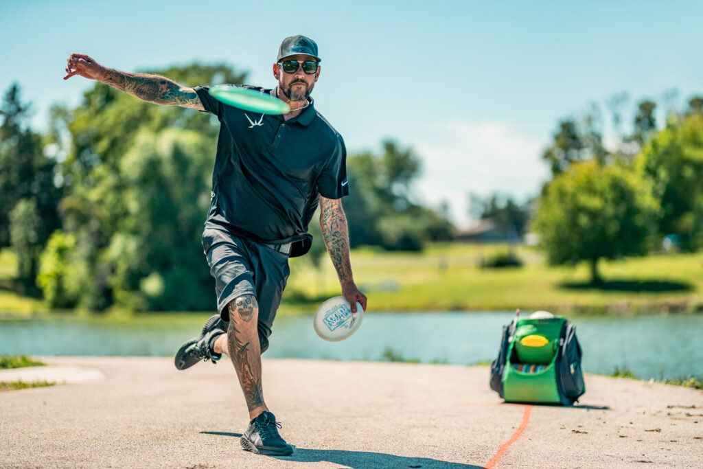 A front-facing view of Patrick Brown as he throws a tee shot off the tee pad. His GRIPeq disc golf bag is sitting on the ground behind him.