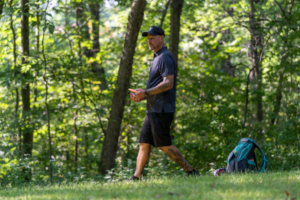 Patrick Brown walks across the course with a disc in-hand. His turquoise GRIPeq AX5 bag sits on the ground behind him.