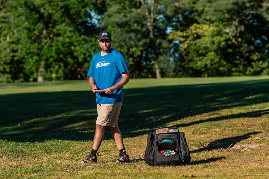 Brian Earhart prepares to throw a disc with a black GRIPeq disc golf bag at his feet. The front flap is open showing his discs inside.
