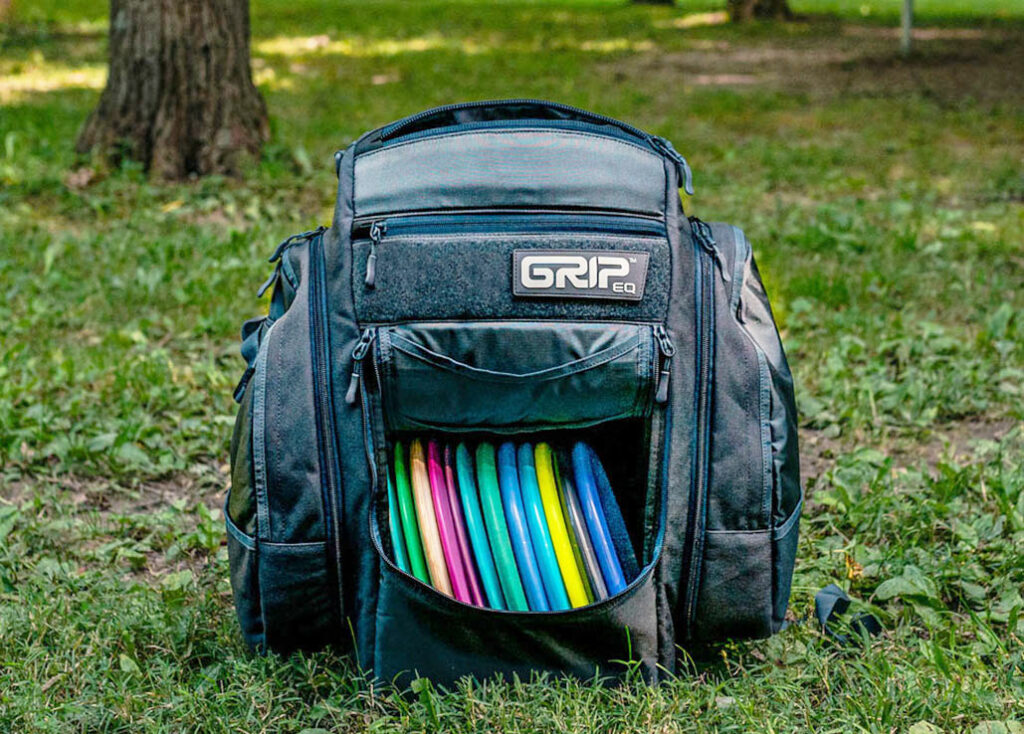 A gray GRIPeq BX3 bag sits in the grass. The front flap is open and the discs inside are visible. There is a GRIPeq velcro patch on the front.