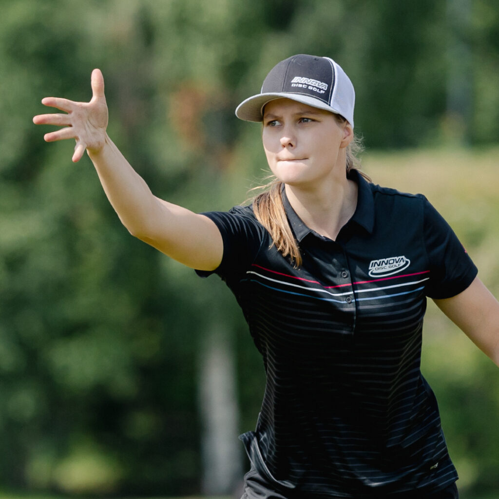 Henna Blomroos, with an outstretched arm and fingers after just releasing a putted disc. She is looking in the direction of the putt.