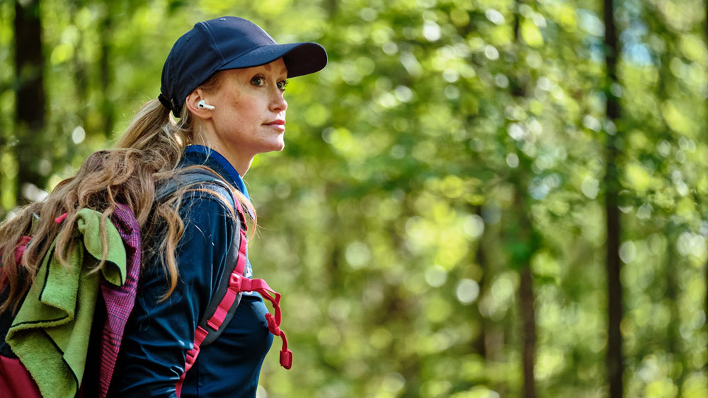 A side view of Holly Finley. She's looking to the right side of the frame and has a baseball hat and earbuds in. She is wearing a pink GRIPeq disc golf bag.
