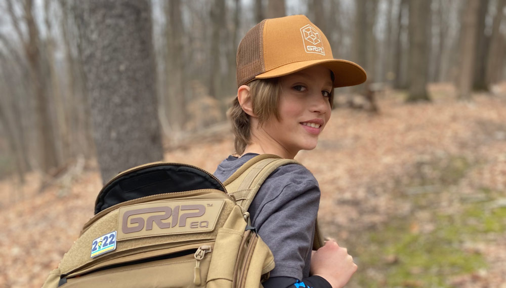 Leighton Brinster looking over his right shoulder at the camera. He's wearing a sand GRIPeq AX5 bag and a sand GRIPeq trucker hat.