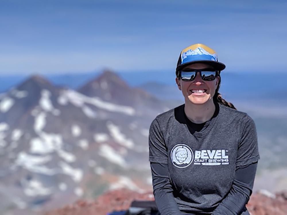Team GRIPeq member, Valarie Jenkins, smiling and facing the camera. She is sitting on top of a mountain with snow covered mountains in the background.