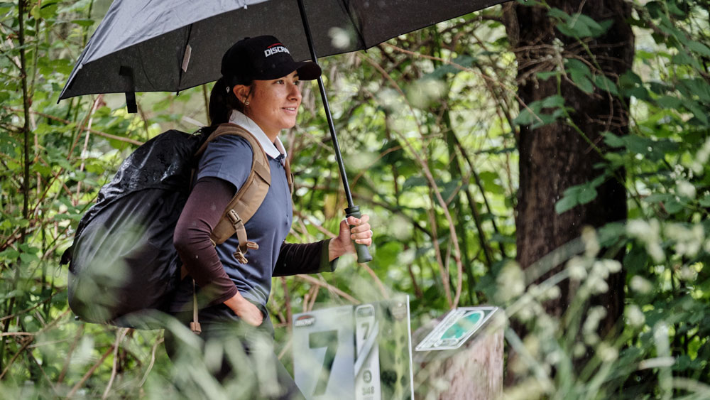 Alexis Mandujano holds an umbrella in the rain during a round. On her back is her sand GRIPeq bag with a GRIPeq rain cover on it.