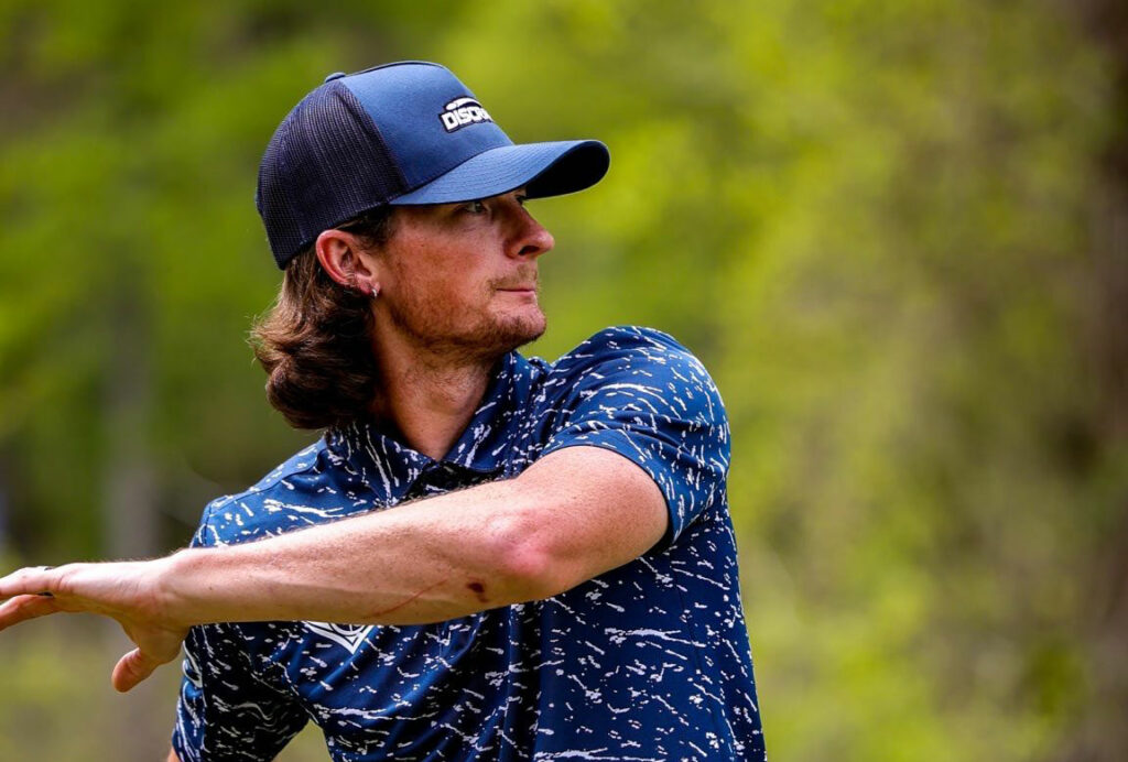 Ben Callaway wearing a Discraft baseball hat with his arm across his torso after just releasing a disc.