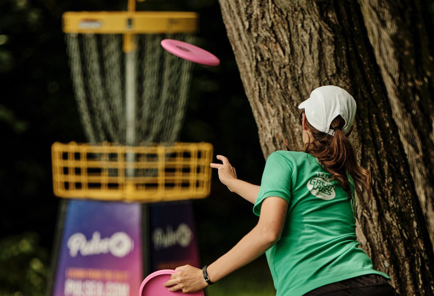 A view of Team GRIPeq member Ali Smith from the back as she releases a disc to putt into a basket in the background.
