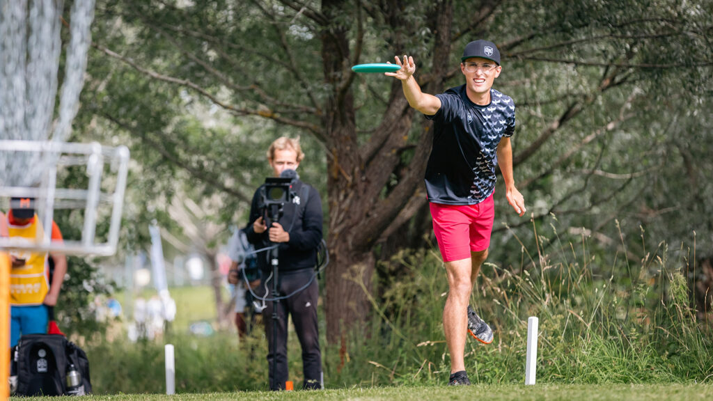 Eagle McMahon releases a putt to throw a disc into a disc golf basket in the foreground.