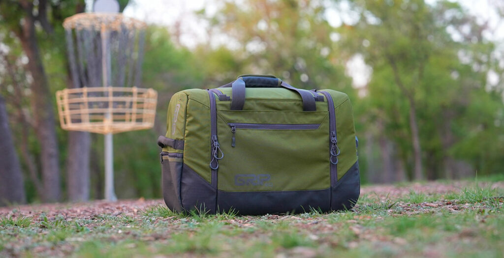 A moss green TSD disc golf bag sits on the ground in front of a disc golf basket.