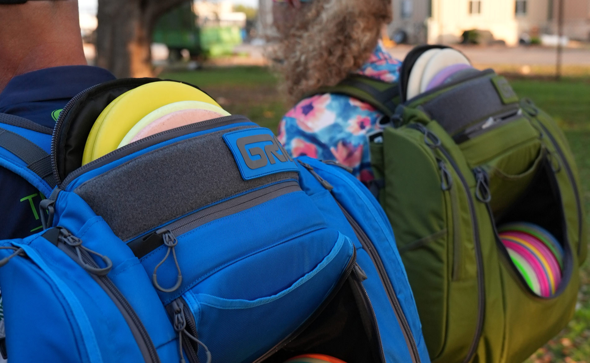 A view from behind of two GRIPeq AX6 bags on the backs of two players. The front pocket is open and there are discs inside as well as in the top putter pocket.